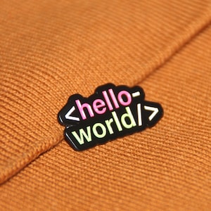 Pode incluir: Um pin preto esmaltado com o texto "hello world" em rosa e verde, com colchetes. O pin está sobre um fundo de tecido laranja tricotado. Um acessório divertido para programadores.