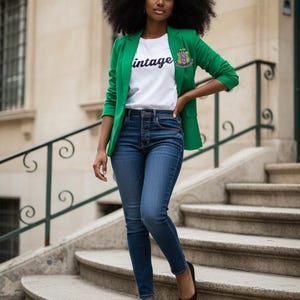 May include: A woman wearing a green blazer with a white t-shirt that says "vintage". She is also wearing blue jeans and black flats. The blazer has a crest on the left side. She is standing on a set of stone stairs.