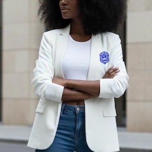 May include: A white blazer with a blue emblem on the lapel, paired with a white top and blue jeans. The blazer has a classic cut and is worn with the arms crossed. The background is a neutral-toned building.