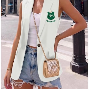 May include: A white sleeveless blazer with a single-button closure, paired with denim shorts. The blazer features a green and white emblem. A small, gold quilted handbag hangs from a chain.