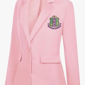 May include: A light pink blazer with a single-button closure and a notched lapel. The blazer features a patch with the letters AKA and the words "DEDICATED TO SERVICE". The brand name "MINT LIMIT" is printed on the collar.