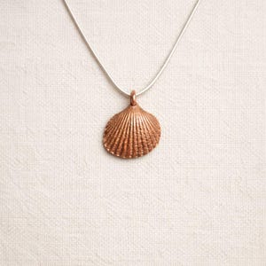 Op de afbeelding: Een koperkleurige schelpenhanger hangt aan een zilveren ketting. De hanger is een gedetailleerde replica van een sint-jakobsschelp, met getextureerde ribbels. De ketting wordt gepresenteerd tegen een neutrale, gebroken witte achtergrond.