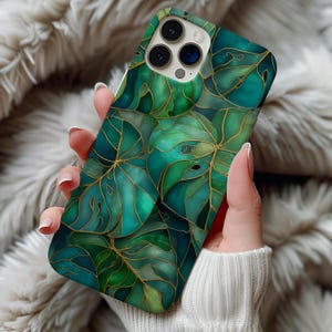 Può includere: Custodia per smartphone con un design di foglie verdi stilizzate con contorni dorati. Le foglie hanno diverse sfumature di verde e blu, creando un effetto a strati. La custodia ha una finitura lucida e un ritaglio per la fotocamera.