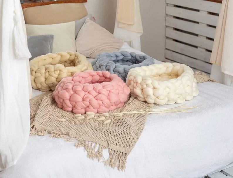 May include: Four knitted pet beds in different colors: beige, pink, white, and blue. The beds are on a white bedspread with a beige knitted throw blanket.