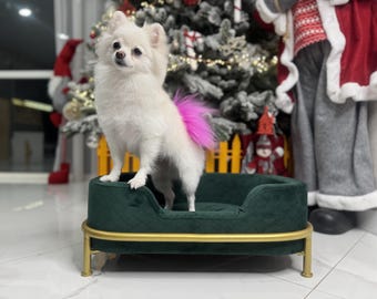 May include: A white fluffy dog with a pink tail sits on a green velvet dog bed with a gold metal frame.