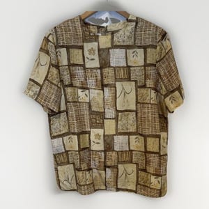May include: A short-sleeved blouse with a patchwork design in shades of brown, beige, and cream. The pattern includes squares with floral and woven textures. The blouse has a round neckline and is hanging on a wooden hanger.
