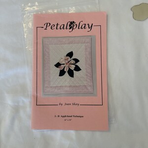 Op de afbeelding: Een roze boekje getiteld "Petal play" met een bloemmotief. Het boekje bevat de tekst "by Joan Shay" en "3-D Appli-bond Technique". De afbeelding toont een gevierkant quilt met een bloemmotief, 35,56 cm x 40,64 cm.