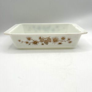 May include: A rectangular white glass baking dish with integrated handles on the short sides. The exterior features a brown printed pattern of pine branches and pinecones centered along the long side of the dish, providing a clear view.