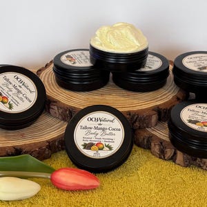 Könnte beinhalten: Mehrere schwarze, runde Dosen mit OCHNatural Tallow-Mango-Cocoa Körperbutter und cremefarbenen Etiketten mit Mango- und Kakaoabbildungen. Eine geöffnete Dose zeigt hellgelbe Körperbutter. Die Etiketten geben 57,5g an.