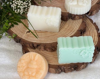 May include: Five handmade soap bars with various designs. The soaps are on a wooden surface. The soaps are white, pink, green, and orange. The soaps have floral and animal designs.