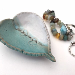 May include: A heart-shaped ceramic dish with a teal and white glaze. The dish has a textured edge and is decorated with small heart shapes. Two silver rings are sitting on a white surface next to the dish.