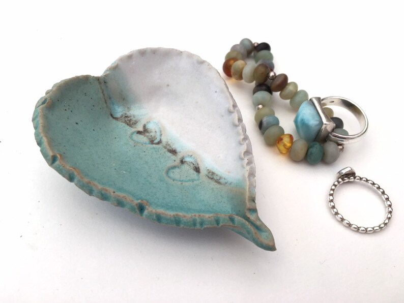 May include: A heart-shaped ceramic dish with a teal and white glaze. The dish has a textured edge and is decorated with small heart shapes. Two silver rings are sitting on a white surface next to the dish.