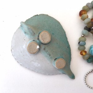 May include: A ceramic dish with a blue and white glaze, featuring three small feet on the bottom. The dish is shaped like a leaf and has a pointed tip. The dish is next to a beaded bracelet with amber and blue stones, and a silver ring with a blue stone.