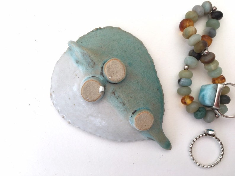 May include: A ceramic dish with a blue and white glaze, featuring three small feet on the bottom. The dish is shaped like a leaf and has a pointed tip. The dish is next to a beaded bracelet with amber and blue stones, and a silver ring with a blue stone.