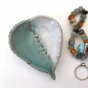 May include: A heart-shaped ceramic dish with a teal and white glaze. The dish has three heart-shaped impressions on the teal side.  A silver ring with a blue stone and a beaded bracelet with amber and turquoise stones are next to the dish.