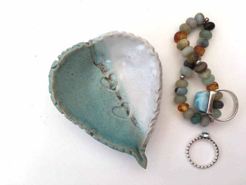 May include: A heart-shaped ceramic dish with a teal and white glaze. The dish has three heart-shaped impressions on the teal side.  A silver ring with a blue stone and a beaded bracelet with amber and turquoise stones are next to the dish.