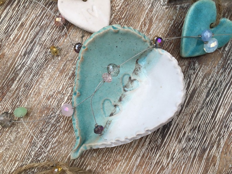 May include: A string of iridescent beads hangs between two ceramic hearts. One heart is white with a blue glaze on the top half, and the other is a solid blue.
