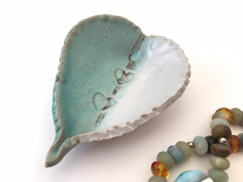 May include: A heart-shaped ceramic dish with a light blue and white glaze. The dish has a rough, textured edge and three small hearts embossed on the surface.