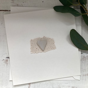 May include: A white card with a small, white heart-shaped paper cutout on a white background. The card is surrounded by green leaves.
