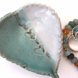 May include: A heart-shaped ceramic dish with a teal and white glaze. The dish has three heart-shaped impressions on the inside. The dish is next to a beaded bracelet with amber and turquoise beads.