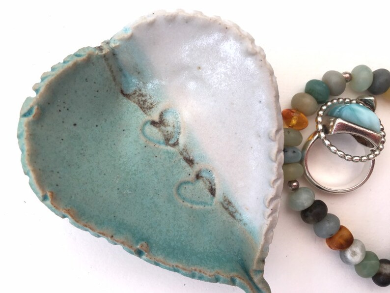 May include: A heart-shaped ceramic dish with a teal and white glaze. The dish has three heart-shaped impressions on the inside. The dish is next to a beaded bracelet with amber and turquoise beads.