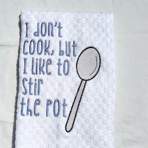 May include: White kitchen towel with blue embroidery that reads "I don't cook but I like to stir the pot" and a silver spoon illustration.