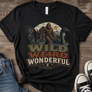 May include: A black short-sleeved t-shirt featuring a graphic print of Bigfoot, a moth, and a sea creature in a forest setting. The text "WILD WEIRD WONDERFUL" is printed below the illustration, accompanied by a small footprint and two crossed arrows.