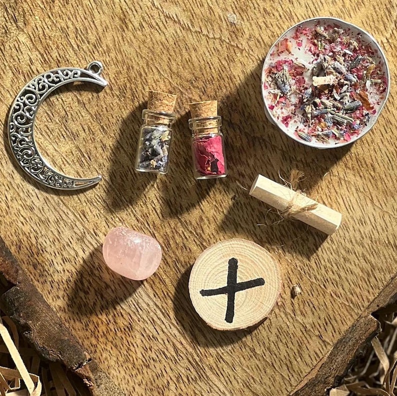 May include: A collection of witchy items including a silver crescent moon charm, two small glass bottles with cork tops filled with dried flowers, a pink crystal, a small wooden disc with a black cross painted on it, a rolled up piece of parchment tied with twine, and a small white candle with dried flowers and herbs inside.