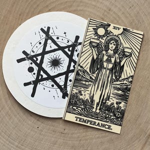 Peut inclure: Une carte de tarot noire et blanche avec le mot "TEMPERANCE" et un motif circulaire avec une &eacute;toile et des &eacute;l&eacute;ments c&eacute;lestes. La carte repr&eacute;sente un ange tenant deux bols. Les articles sont sur une surface en bois.