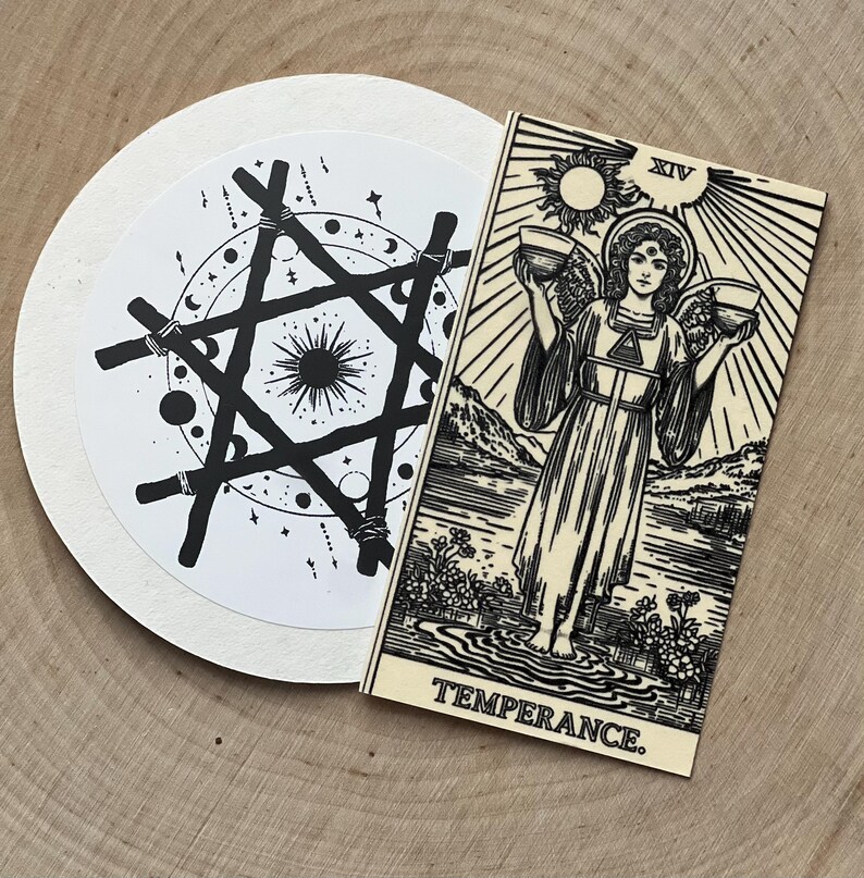 Peut inclure: Une carte de tarot noire et blanche avec le mot "TEMPERANCE" et un motif circulaire avec une &eacute;toile et des &eacute;l&eacute;ments c&eacute;lestes. La carte repr&eacute;sente un ange tenant deux bols. Les articles sont sur une surface en bois.