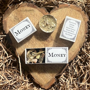 Pode incluir: Três pequenas caixas brancas com a palavra "MONEY" impressa nelas. As caixas contêm um amuleto de chave, uma pequena garrafa de ervas e uma vela branca com ervas secas.