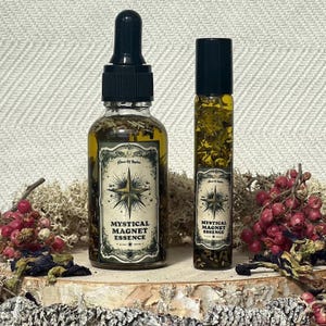 May include: Two bottles of Mystical Magnet Essence oil, one in a dropper bottle and one in a roller bottle. Both bottles are clear glass and contain a yellow liquid with herbs and flowers inside. The bottles have a black label with a gold star and the text "Mystical Magnet Essence".