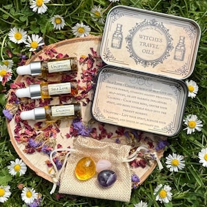 Peut inclure: Un ensemble de trois petites bouteilles en verre avec des étiquettes indiquant "Uplifting", "Self Love" et "Calming". Les bouteilles sont dans une boîte en métal avec l'inscription "Witches Travel Oils" sur le couvercle. La boîte est ouverte et il y a une petite pochette avec trois cristaux à l'intérieur.