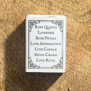 May include: A white box with black text that reads "Rose Quartz Lavender Rose Petals Love Affirmation Love Candle Moon Charm Love Rune".
