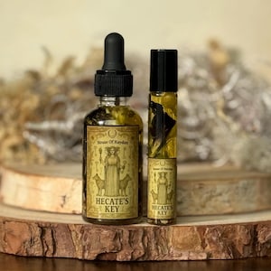 May include: Two glass bottles of essential oil, one with a dropper and the other a rollerball, sit on a wooden surface. The labels read "House of Raydan" and "Hecate's Key" with an illustration of a woman and dogs. The oils are amber-colored.