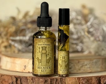 Pode incluir: Duas garrafas de vidro de óleo essencial, uma com conta-gotas e outra roll-on, estão sobre uma superfície de madeira. Os rótulos dizem "House of Raydan" e "Hecate's Key" com uma ilustração de uma mulher e cães. Os óleos são de cor âmbar.