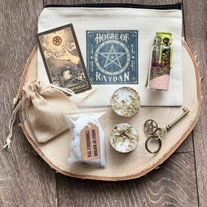 Peut inclure: Une pochette en toile blanche avec le texte "House of Raydan" et une étoile à cinq branches. La pochette contient une carte de tarot, un petit sac en toile de jute, deux bougies chauffe-plat, un petit sachet de camomille, une bouteille en verre avec du sel rose et des herbes, et une clé avec un charme pentagramme.
