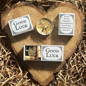 Peut inclure: Une boîte blanche avec les mots "Good Luck" imprimés dessus. La boîte contient une petite bougie, un charme en fer à cheval, une petite bouteille d'herbes et un morceau de papier roulé. La boîte est posée sur une surface en bois.