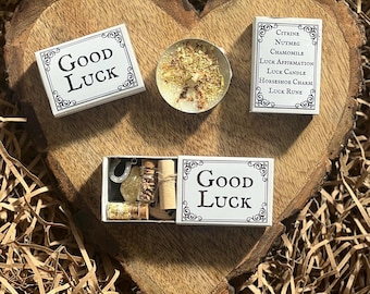 May include: A white box with the words "Good Luck" printed on it. The box contains a small candle, a horseshoe charm, a small bottle of herbs, and a rolled up piece of paper. The box is sitting on a wooden surface.