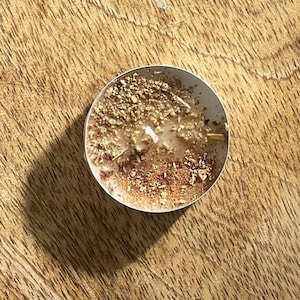 May include: A close-up of a small, round, brown tea light candle with a white wax center. The candle is surrounded by dried herbs and spices.