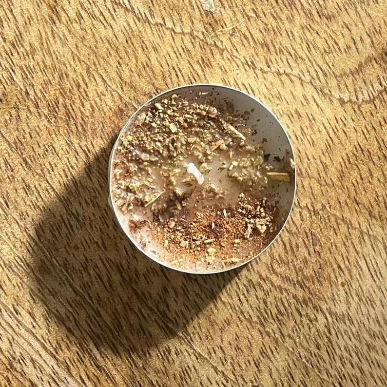 May include: A close-up of a small, round, brown tea light candle with a white wax center. The candle is surrounded by dried herbs and spices.