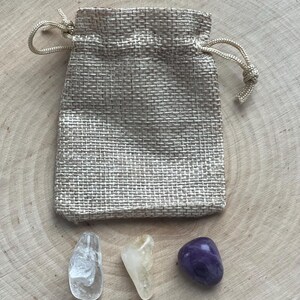 Peut inclure: Un petit sac en toile de jute beige avec une fermeture &agrave; cordon naturelle. Trois pierres pr&eacute;cieuses polies sont expos&eacute;es en dessous : un quartz clair, une citrine jaune et une am&eacute;thyste violet fonc&eacute;. Les pierres sont sur une surface en bois clair.