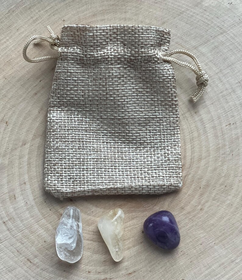 Peut inclure: Un petit sac en toile de jute beige avec une fermeture &agrave; cordon naturelle. Trois pierres pr&eacute;cieuses polies sont expos&eacute;es en dessous : un quartz clair, une citrine jaune et une am&eacute;thyste violet fonc&eacute;. Les pierres sont sur une surface en bois clair.