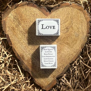 May include: Two small white boxes with black trim and ornate designs. The top box says "LOVE" in black lettering. The bottom box lists "Rose Quartz, Lavender, Rose Petals, Love Affirmation, Love Candle, Moon Charm, Love Rune".