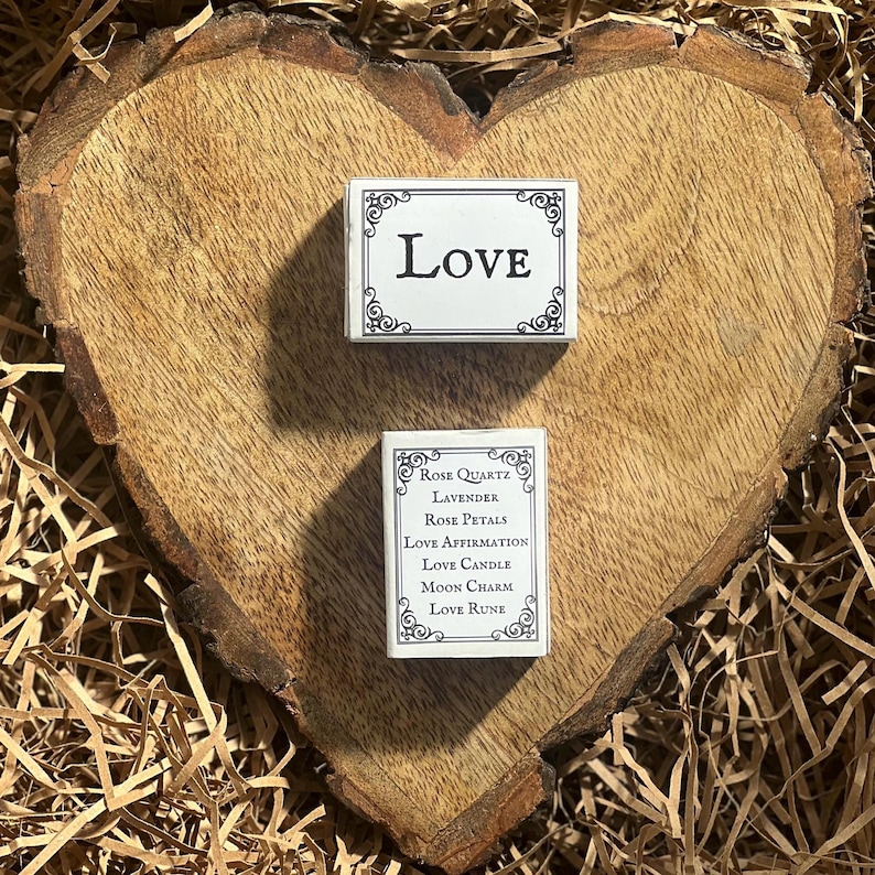 May include: Two small white boxes with black trim and ornate designs. The top box says "LOVE" in black lettering. The bottom box lists "Rose Quartz, Lavender, Rose Petals, Love Affirmation, Love Candle, Moon Charm, Love Rune".