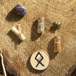 May include: A wooden circle with a black rune symbol, a small glass bottle with a cork top filled with brown powder, a small glass bottle with a cork top filled with tan powder, a small rolled up piece of paper tied with twine, and a purple amethyst stone.