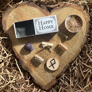 May include: A wooden heart-shaped board with a small box that says "Happy Home", a purple crystal, a small rolled up piece of paper, a small bottle with a cork stopper, a small wooden circle with a rune symbol, a small bottle with a cork stopper, a small candle with a gold lid, and a small gold bell charm.