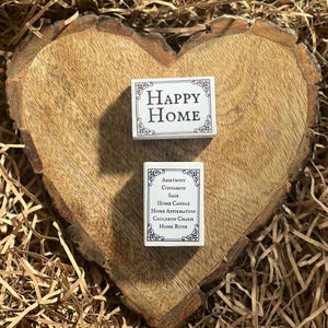 May include: Two white boxes with black text. The top box says "HAPPY HOME". The bottom box says "Amethyst Cinnamon Sage Home Candle Home Affirmation Cauldron Charm Home Rune".