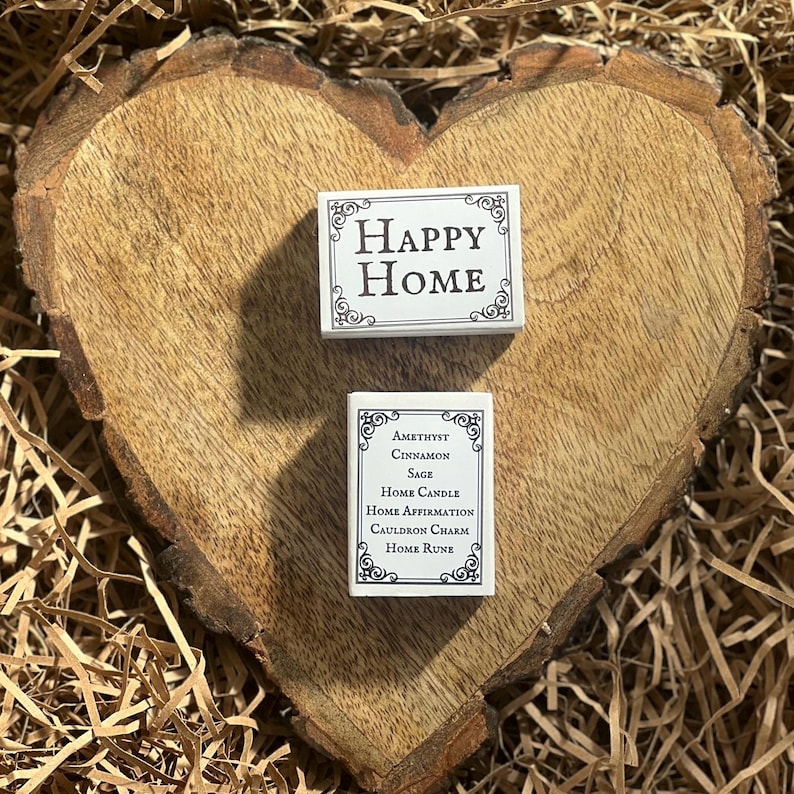 May include: Two white boxes with black text. The top box says "HAPPY HOME". The bottom box says "Amethyst Cinnamon Sage Home Candle Home Affirmation Cauldron Charm Home Rune".
