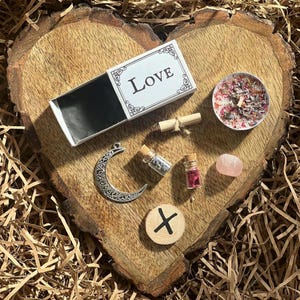 May include: A heart-shaped wooden board with a variety of love-themed items, including a matchbox labeled "Love", a small candle, a crescent moon charm, a small bottle of herbs, a small bottle of crystals, a pink stone, and a wooden rune.